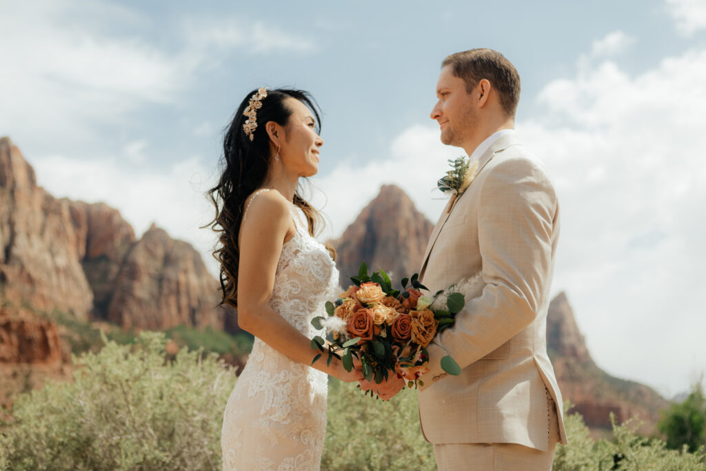 Rina and Derek facing each other with a bouquet of peach roses beneath the red cliffs of Zion at their vow renewal