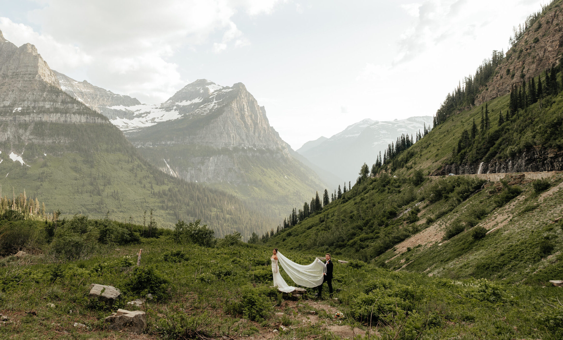 Ashley and Caleb holding her long veil in a wide green valley among the peaks of Glacier National Park