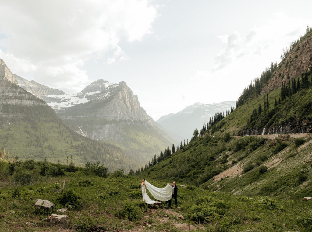Ashley and Caleb holding her long veil in a wide green valley among the peaks of Glacier National Park