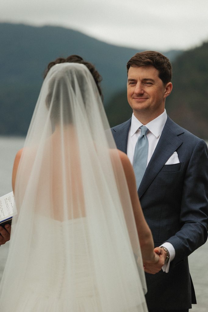 Jamie smiling at Beth through her sheer veil as they exchange vows by the lake