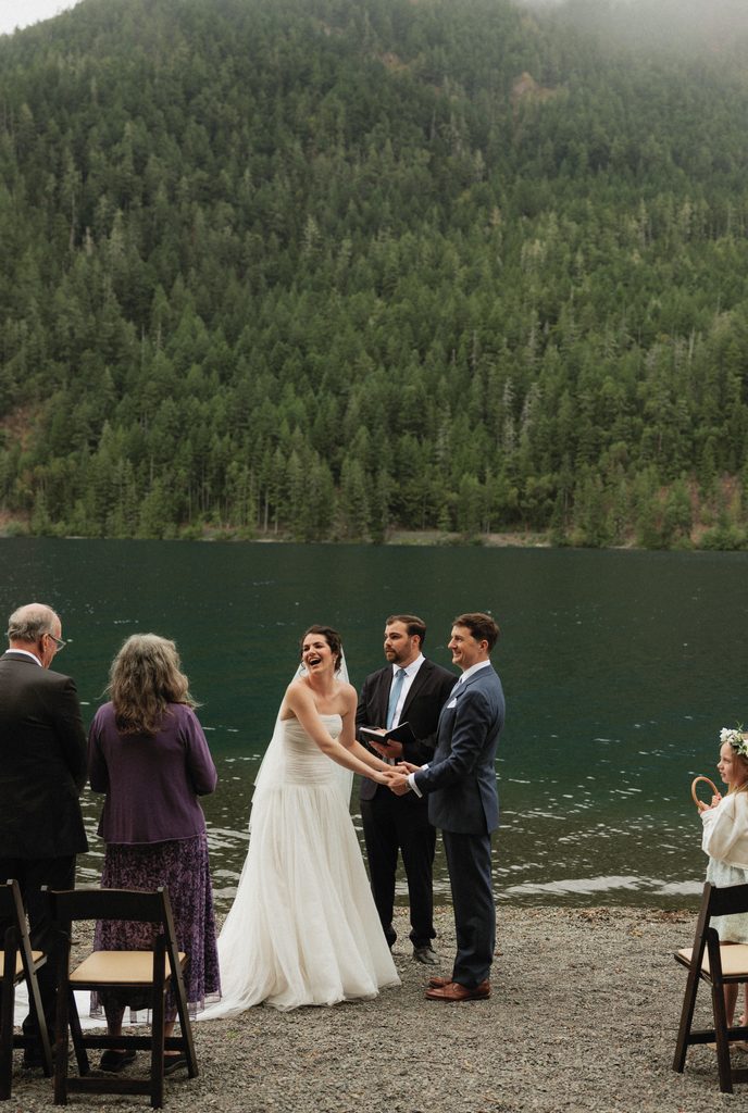 Beth and Jamie hold hands before their officiant and loved ones on the shore of Lake Crescent