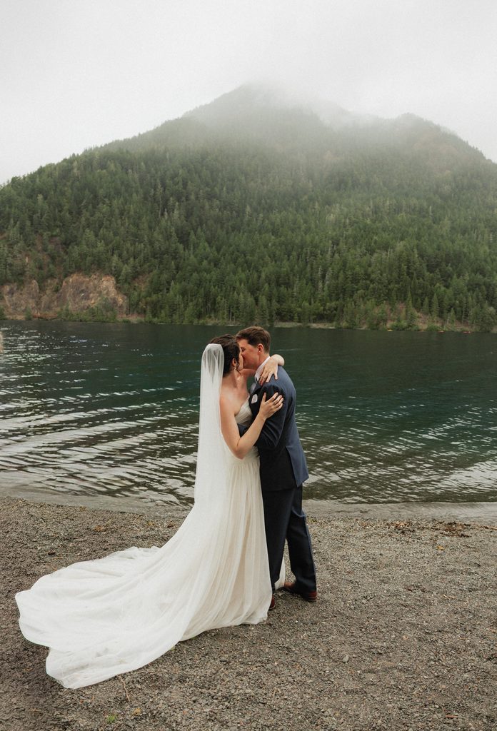 Beth and Jamie kissing on the pebbled shore of Lake Crescent as her long train spreads behind her