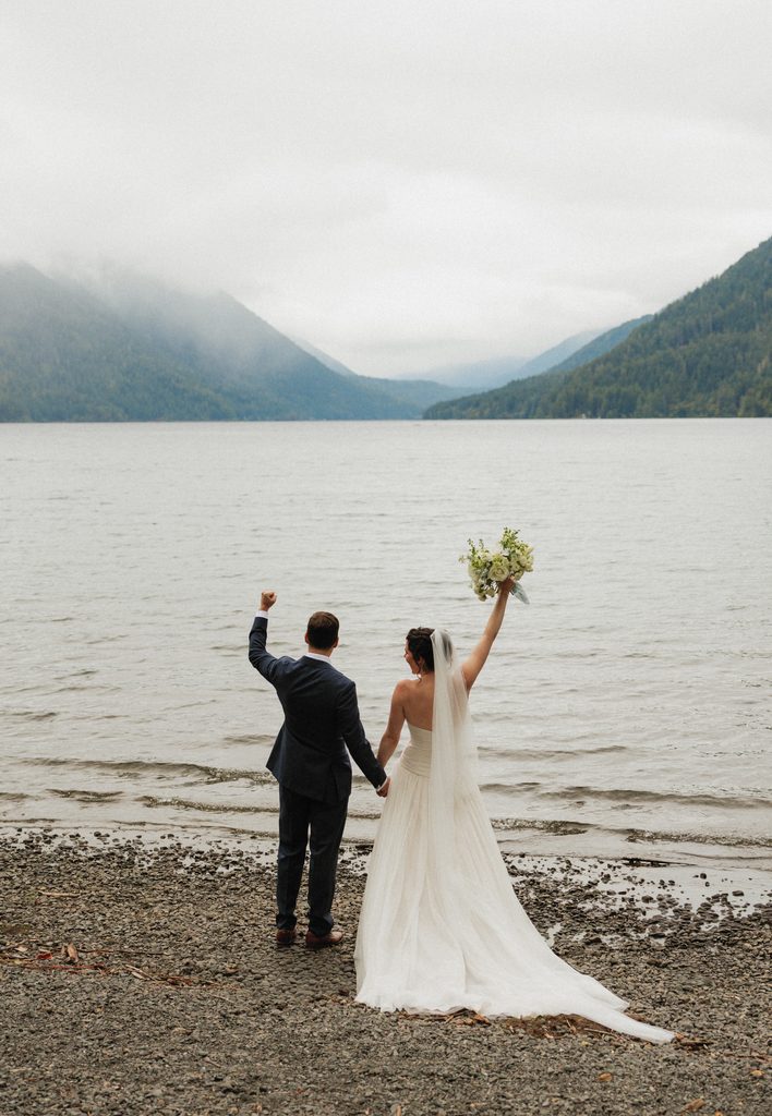 Beth and Jamie cheer with her bouquet raised high on the shore of Lake Crescent
