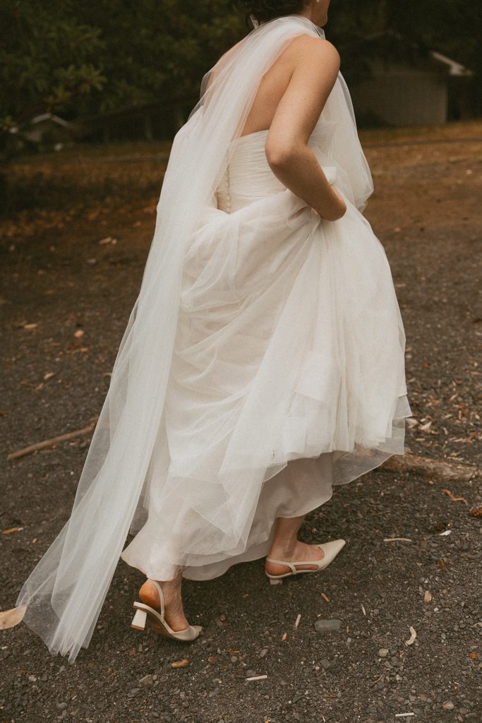 Beth lifts her gown and veil as she walks across a gravel path in strappy heels