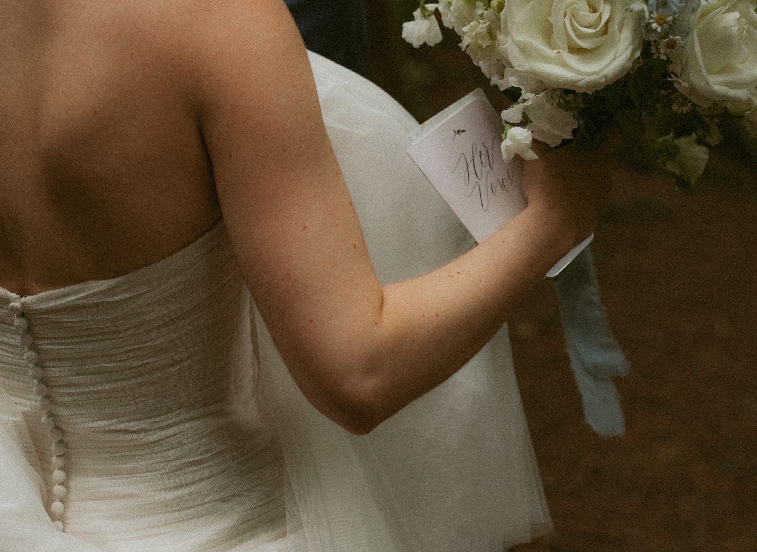 Bride's buttoned gown seen from behind as she holds her vow book and a bouquet of white roses