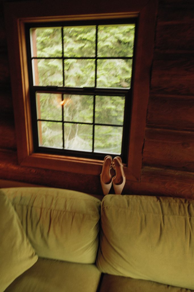 Bridal heels resting on a window sill inside a log cabin with green trees outside