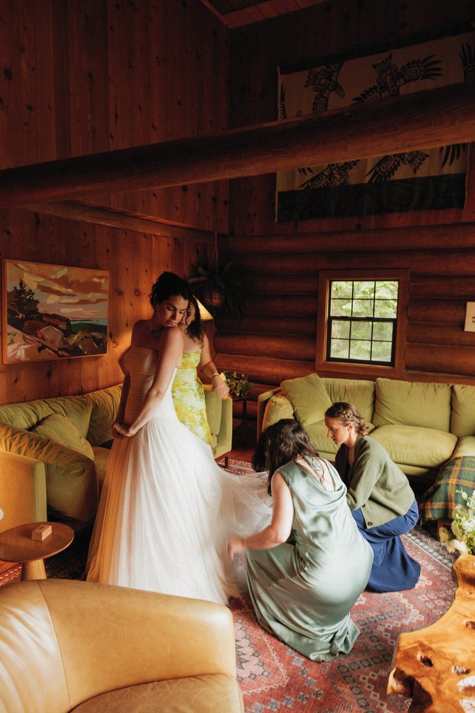 Bridesmaids in sage green help Beth into her gown inside a warm wooden cabin