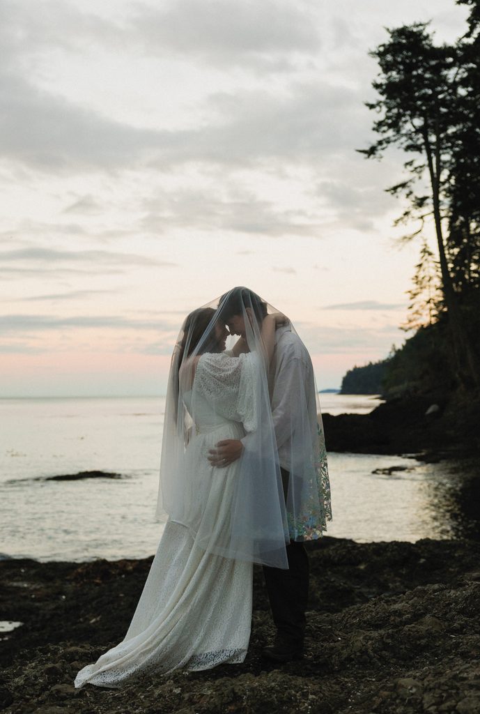Dakota and Rowdy embracing under her veil on a quiet beach at dusk