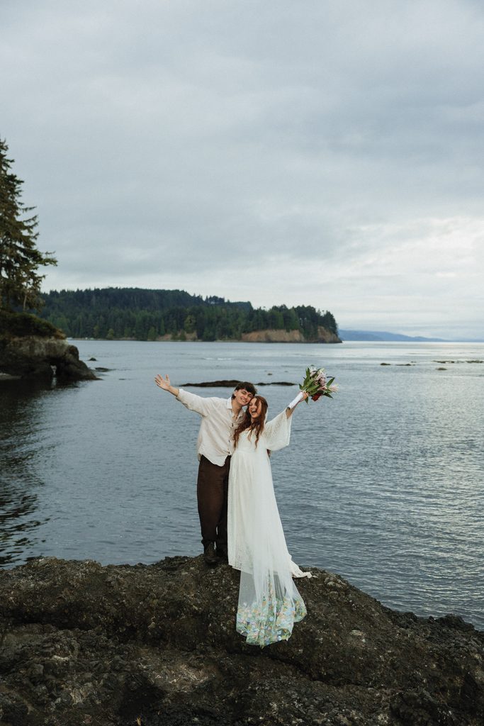 Dakota and Rowdy raise their arms in celebration on a rocky point above calm Pacific Northwest water