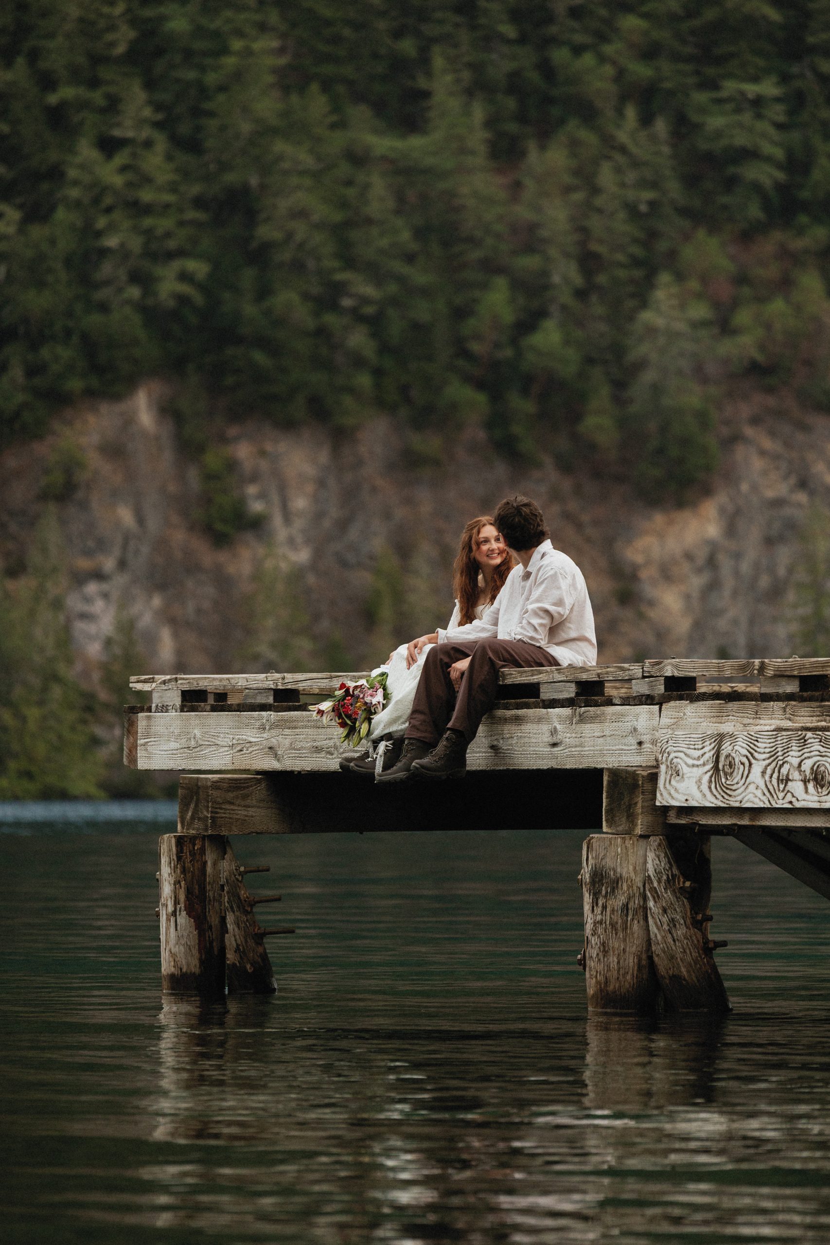 Dakota and Rowdy sit close together on an old wooden pier over Lake Crescent