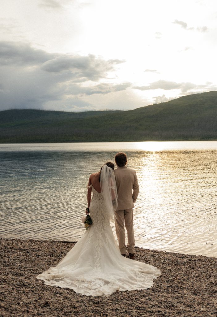 Bride and groom seen from behind on the pebbled shore of Lake McDonald as evening light hits the water