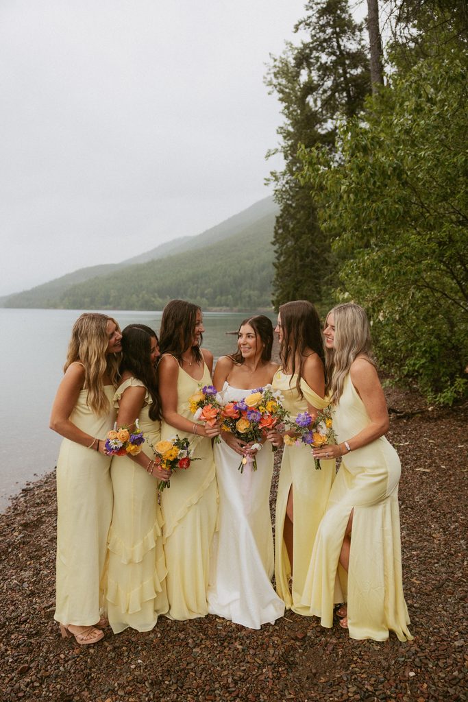 Sydney laughing with her bridesmaids in pale yellow dresses on a Glacier lakeshore beneath forested mountains
