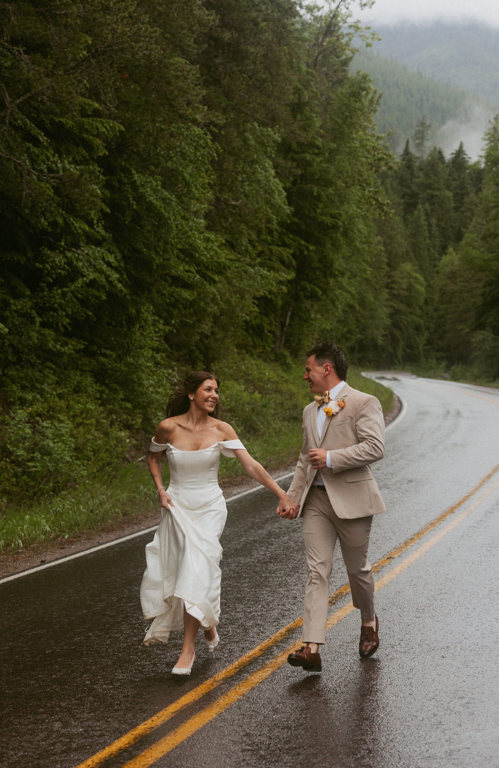 Sydney and Austin running hand in hand down a rain-soaked road through misty evergreen forest