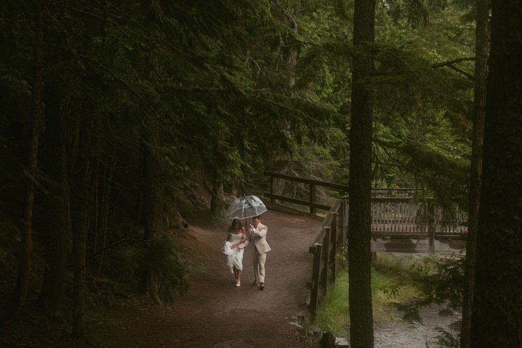 Sydney and Austin walk hand in hand under an umbrella along a misty forest trail in Glacier National Park