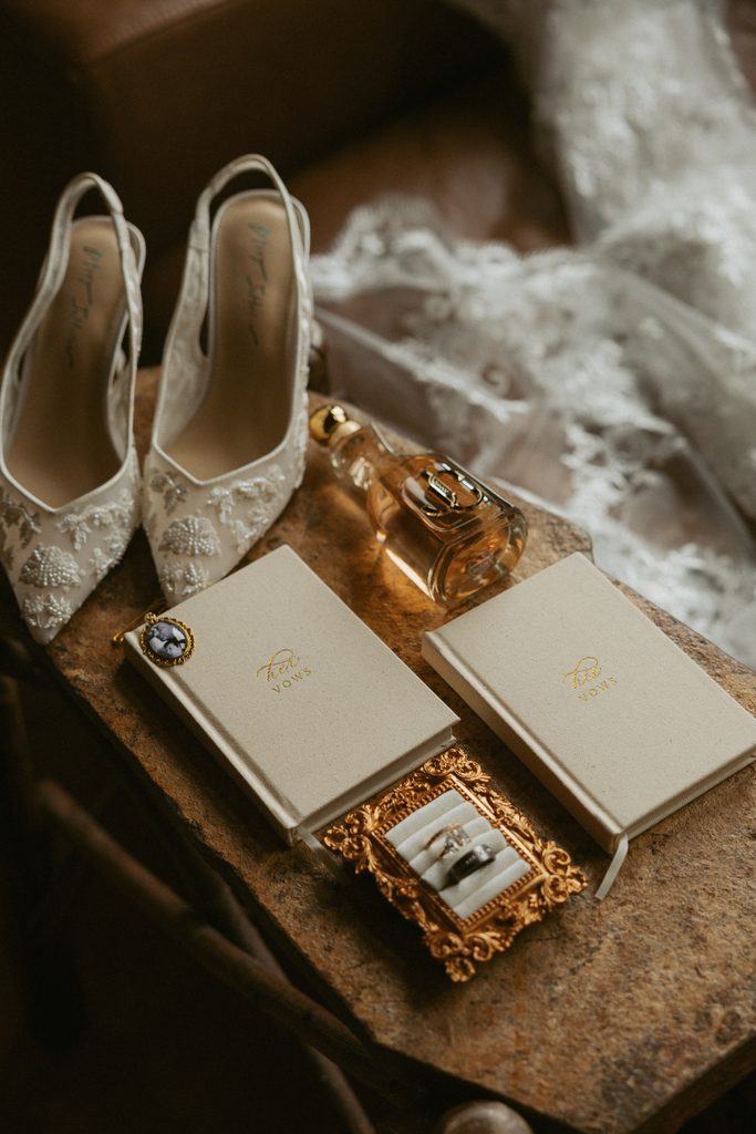 Lace bridal heels, a perfume bottle, a ring box and two vow books laid out on a wooden table