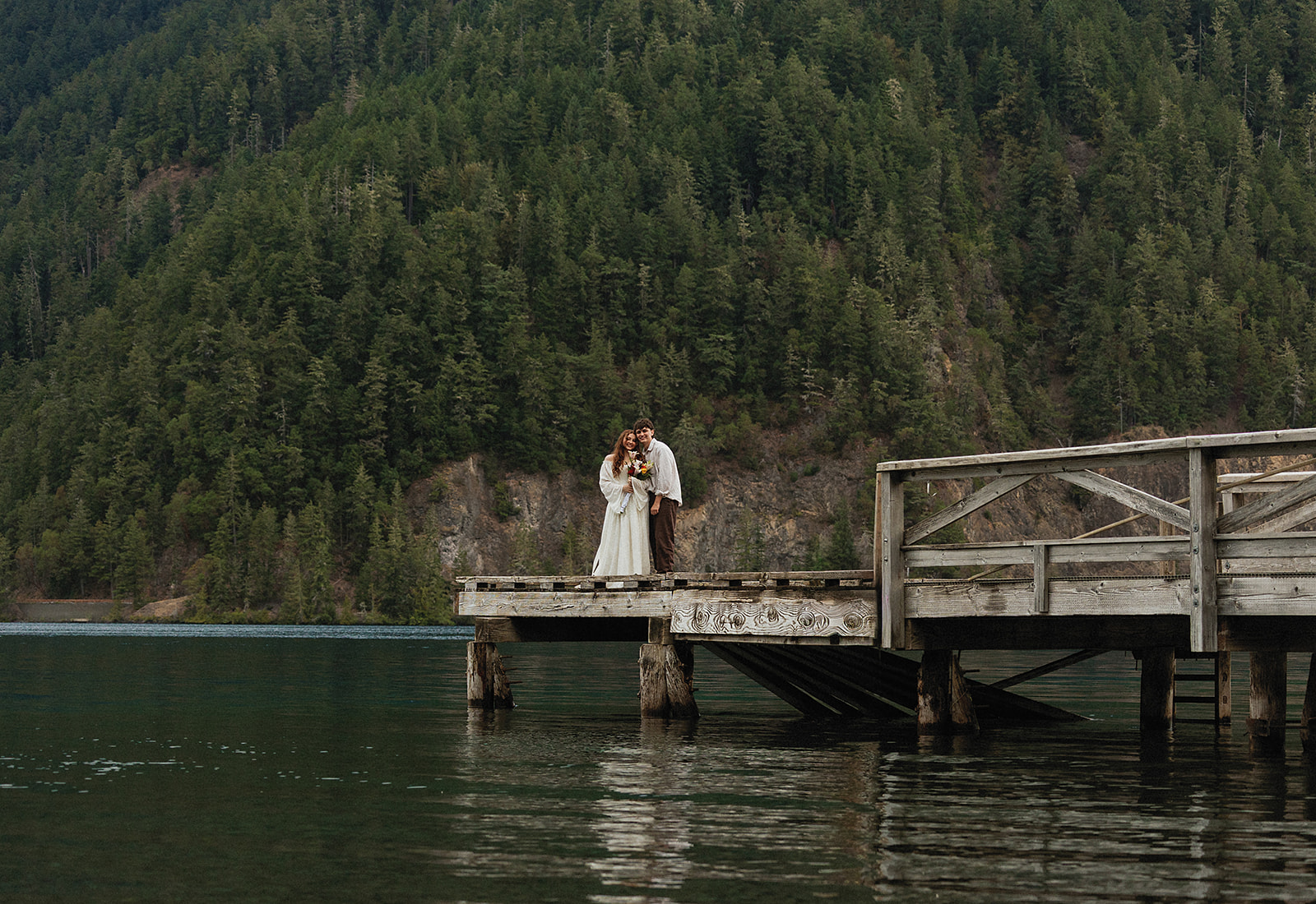 Dakota and Rowdy stand on a wooden dock reaching out over Lake Crescent, surrounded by forested hills